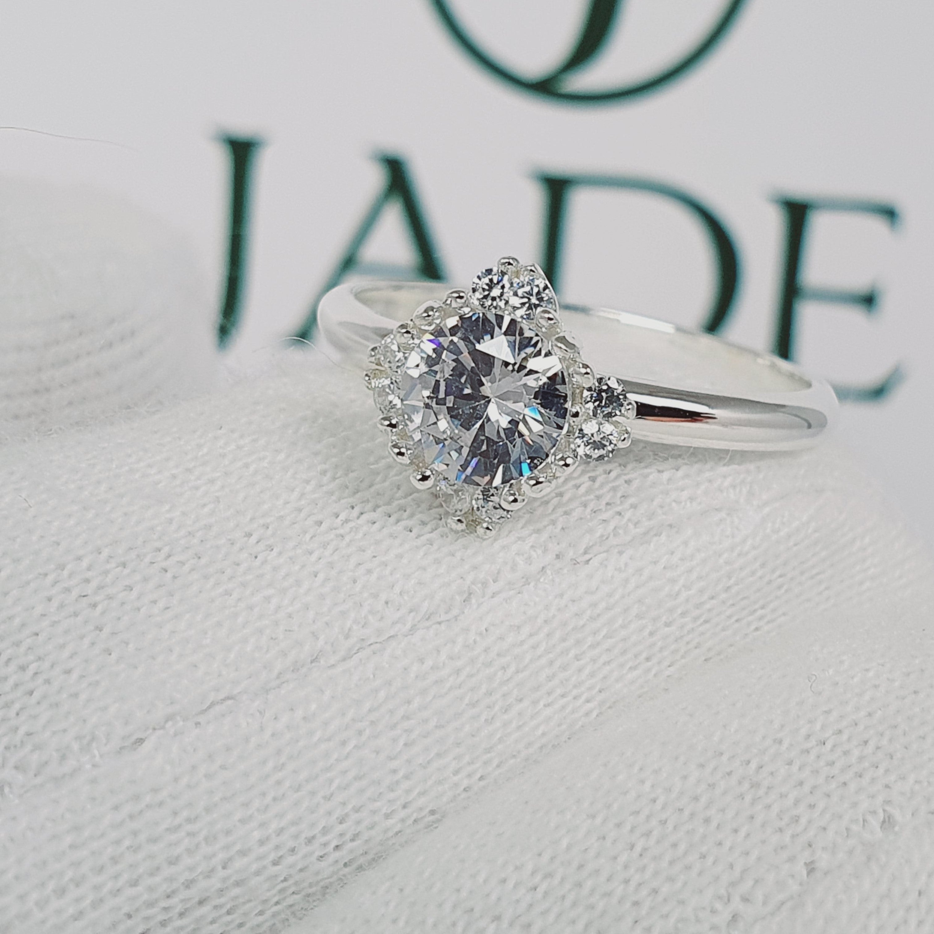 Jade18k Damelia engagement ring in silver with a central round-cut diamond surrounded by smaller stones in a floral halo setting.